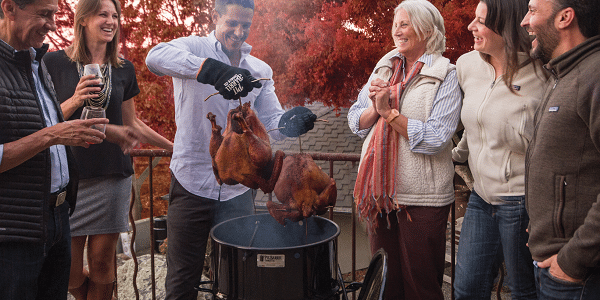 Friends on an outdoor deck standing around a barrel smoker, laughing and drinking wine. A man lifts two smoked turkeys out of the smoker.